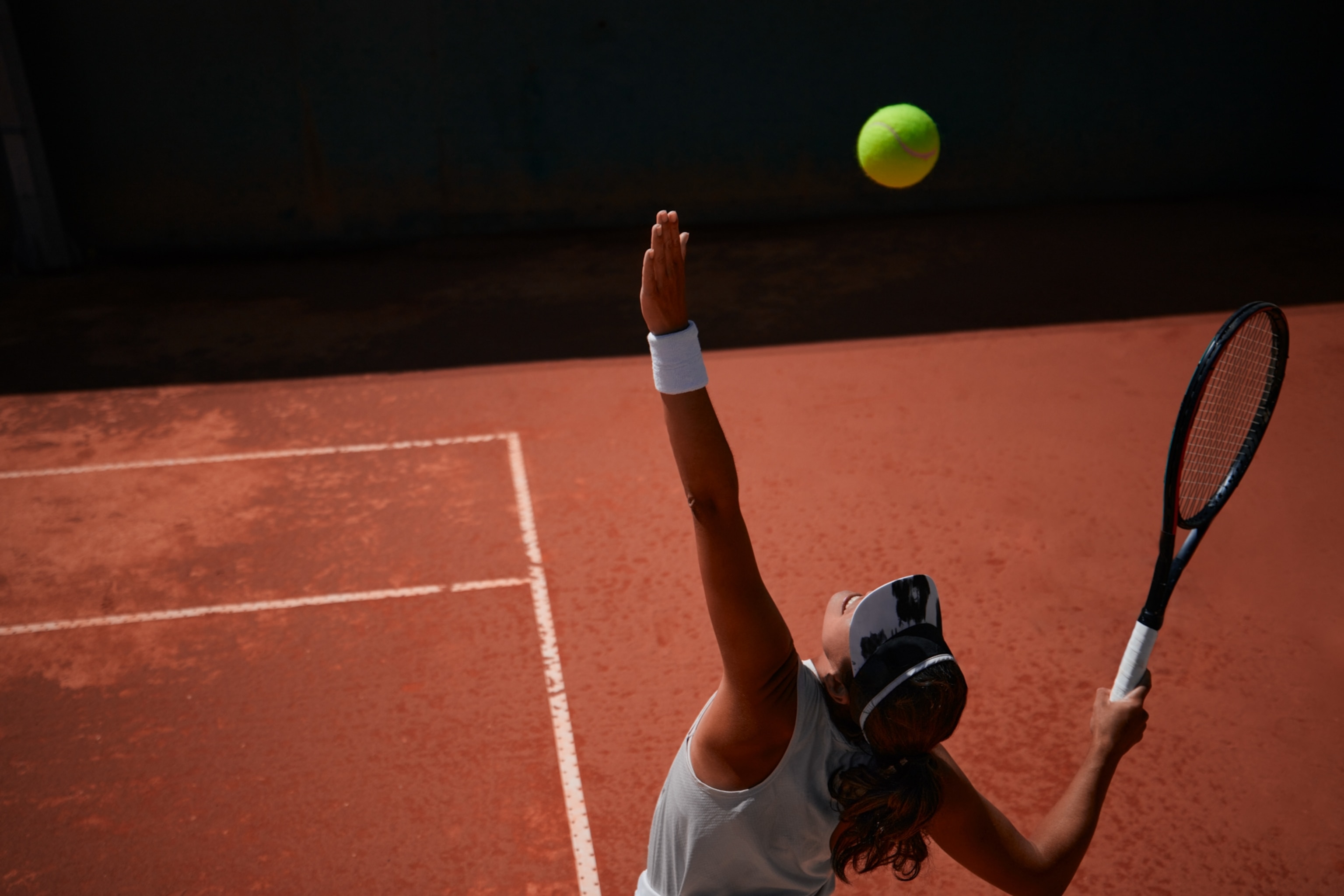 The upper body of a tennis player mid-serve, her serving arm and head turned upward as she tracks the tennis ball, which is seen at the top of the image.