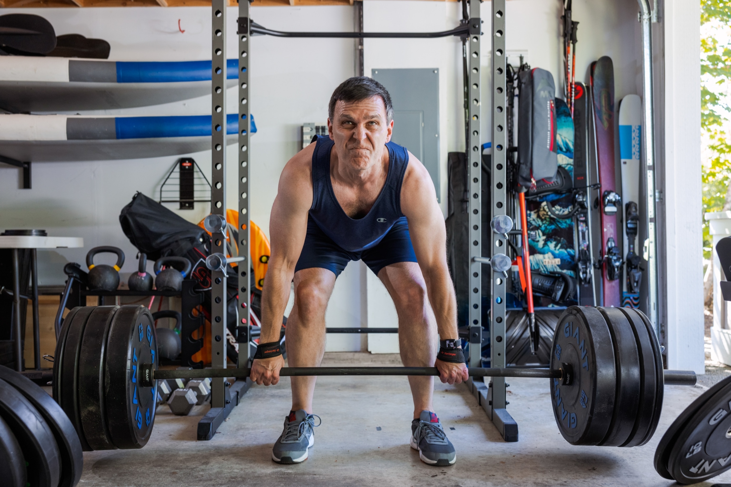 A middle-aged man, bent over and facing the camera as he deadlifts over 300 pounds in his garage.